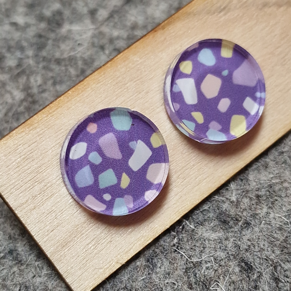 Two round acrylic studs with a purple terrazzo pattern, featuring white, pink, and light blue chips, displayed on a wooden block over a gray felt background.